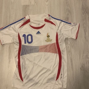 Frankrike Zidane 10 fotbollströja Adidas - Säljer en klassisk fransk landslagströja från Adidas med Zidane och nummer 10 på ryggen. Tröjan är vit med blå och röda detaljer, korta ärmar och tryck på både fram- och baksida. Perfekt för dig som älskar fotbollshistoria och snygg retrostil.