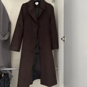 Brun lång kappa från Kappahl - Snygg lång kappa i mörkbrunt från Kappahl, med klassisk krage och knäppning framtill. Kappan har ett bälte i midjan för extra stilpoäng och är tillverkad i mjukt ullmaterial. Perfekt för dig som gillar en clean och tidlös look under höst och vinter.