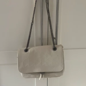 Beige mönstrad axelväska med kedja - Snygg beige axelväska med quiltad design och silverfärgad kedjerem. Väskan är endast använd 4 gånger. 