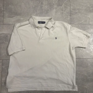 Vit pikétröja från Ralph Lauren - Klassisk vit pikétröja från Ralph Lauren med korta ärmar och krage. Tröjan har två knappar vid halsen och den ikoniska broderade loggan i marinblått på bröstet. Perfekt för en stilren och avslappnad look.