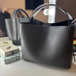 Svart stilren axelväska  - Snygg svart axelväska i slätt skinn. ett stort huvudfack med innerficka som stängs med dragkedja. Perfekt för dig som gillar stilrena accessoarer.