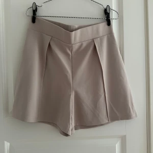 Beige högmidjade shorts från H&M - Stilrena beige shorts från H&M med hög midja och snyggt vikt linning. De har en lätt utsvängd passform och är tillverkade i mjuk polyester. Perfekta för en clean och trendig look under varmare dagar.
