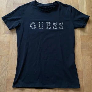 Svart t-shirt från Guess med strass - Svart t-shirt från Guess med korta ärmar och rund hals. Framsidan har GUESS-loggan i glittriga strasstenar som ger en snygg touch. Skön passform och tillverkad i mjuk bomull, perfekt till jeans eller kjol. Är liten i storleken. Passar både XS och S. 