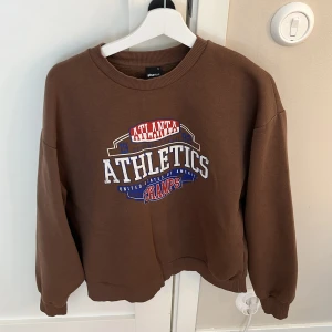 Brun sweatshirt från Gina Tricot - Cool brun sweatshirt från Gina Tricot i storlek S med tryck i vitt, blått och rött där det står 'Atlanta Athletics Champs'. Tröjan har rund hals, långa ärmar och loose passform. Perfekt för dig som gillar en avslappnad stil. Själv stylar jag den med ett par mörkblåa jeans!❤️