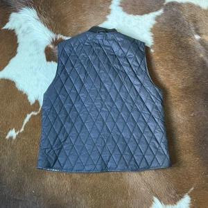 Svart quiltad väst från Gant - Quiltad svart väst från Gant med dragkedja framtill och ribbad krage. Utsidan är stilren med diamantmönster, medan insidan har ett blåvitt rutigt foder. Storleken är XL men passar mer för L.Tveka inte på att höra av er vid frågor!
