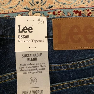 Lee Oscar Relaxed Tapered jeans blå - Snygga blå jeans från Lee i modellen Oscar Relaxed Tapered. Jeansen har klassisk femficksdesign, gul kontrastsöm och är tillverkade i ett hållbart material med minst 20% alternativa fibrer. Passformen är relaxed med avsmalnande ben.