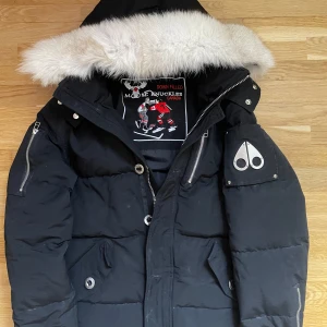 Svart dunjacka Moose Knuckles - Säljer en svart dunjacka från Moose Knuckles med stor huva och vit päls. Jackan har dragkedjor på ärmarna, stora knappar och logga på ärmen. Fodret har broderat motiv med hockeyspelare. Perfekt för kalla dagar.