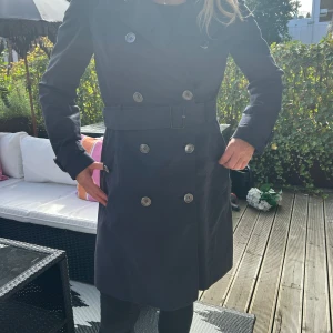 Svart trenchcoat från Hugo Boss - Snygg svart trenchcoat från Hugo Boss med klassisk dubbelknäppning, bälte i midjan och detaljer vid ärmslut. Jackan har långa ärmar och stilren krage, perfekt för dig som vill ha en clean och tidlös look. Materialet känns slitstarkt och jackan har en riktigt schysst passform.
