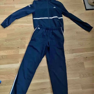 Mörkblå träningsbyxor från Lacoste - Snygga mörkblå träningsbyxor från Lacoste med vit sidodetalj och elastisk midja. Byxorna har en sportig look och är perfekta för dig som gillar streetwear eller vill ha något bekvämt till gymmet. Klassisk Lacoste-logga vid fickan. Bud inget under 1000