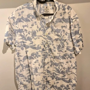 Blommig kortärmad skjorta från DF - Snygg kortärmad skjorta från DF i slim fit-modell. Skjortan är vit med ljusblått blommigt mönster och har knappar framtill samt en bröstficka. Perfekt för dig som gillar en fräsch och somrig stil.