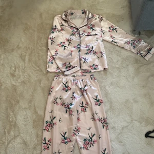 Blommiga pyjamasbyxor och skjorta från SHEIN - Snygga pyjamasbyxor och skjorta från SHEIN i ljusrosa satinliknande material med blommigt mönster i grönt och rosa. Byxorna har elastisk midja och en loose passform, perfekta för chillkvällar hemma.