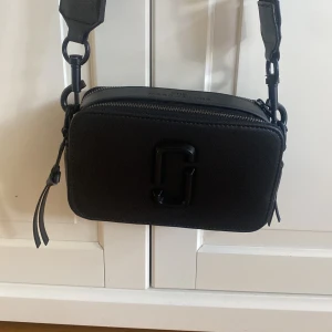 Svart axelväska  - Helt ny svart axelväska likt Marc Jacobs med bred justerbar rem och dubbla dragkedjor. Väskan har en stilren, boxig form och med en diskret logga framtill. Perfekt storlek för det viktigaste och snygg detalj till din outfit.