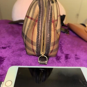 Burberry rutig handväska i skinn -  från Burberry med det ikoniska rutiga mönstret i beige, svart och rött. Väskan är tillverkad i skinn och har en dragkedja upptill. Perfekt storlek för det viktigaste och snygg att bära till alla outfits.