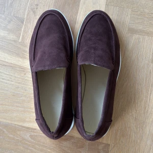 Bruna mockaloafers med vit sula - Snygga loafers i mörkbrun mocka med stilren design och vit platt sula. Skorna har rund tå och är perfekta för dig som gillar en klassisk men avslappnad look. Passar till både jeans och chinos för en clean stil. De är aldrig använda pga för liten storlek. Står 43 på sulan, men stl är troligen 42,5
