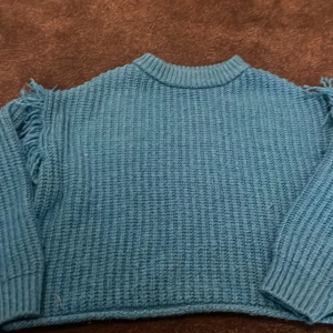 Blå stickad tröja med fransar från Zara - Säljer en blå stickad tröja från Zara med grov struktur och coola fransdetaljer på axlarna. Tröjan har lång ärm, ribbad hals och muddar. Perfekt för dig som gillar en chill och trendig look.