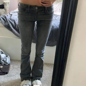 Grå bootcut jeans med kontrastsömmar - Säljer ett par grå jeans med bootcut-modell och snygga kontrastsömmar. Byxorna har låg midja, klassiska fickor bak med lock och dekorativa knappar. Jeansen är i stretchigt material och har en riktigt schysst passform.