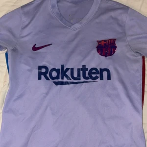 Ljuslila FC Barcelona Nike fotbollströja - Snygg FC Barcelona matchtröja från Nike i ljuslila med mörkblå Rakuten-tryck och klubbmärke i rött och blått. Kortärmad med V-ringning och blå sidopanel. Tillverkad i lätt Dri-FIT material som andas. UNICEF-logga på ryggen. Perfekt för dig som älskar Barca!