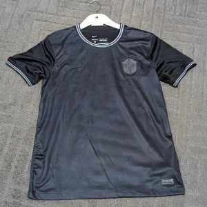Svart Brasil Nike fotbollströja S - Snygg svart Brasilien fotbollströja från Nike med broderat CBF-emblem och fem stjärnor på bröstet. Tröjan har korta ärmar och ribbad krage med grå och vita ränder. Tillverkad i lätt Dri-Fit polyester som andas och håller dig torr under matchen.
