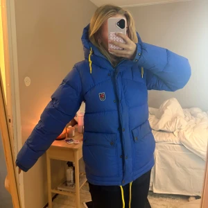 Blå dunjacka från Fjällräven - Säljer den populära blå dunjacka från Fjällräven med huva och gula snören. Det är den nya modellen och inte den som är tjockare! Använd i 2 vintrar så den är lite smutsig, tex lite sminkfläckar vid ”kragen” av jackan, men den går superbra att slänga in i tvättmaskin och även lämna in på kemtvätt om man skulle vilja det!! Verklig i storlek, varken större eller mindre