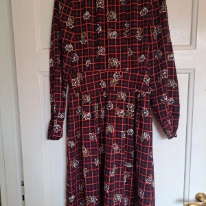 Mönstrad långklänning Holly & Whyte - Långklänning från Holly & Whyte med svart bas, röda rutor och blommigt mönster. Klänningen har långa ärmar med knappar vid ärmslut och markerad midja. Perfekt för dig som gillar unika prints och en klassisk men cool look.