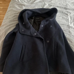 Mörkblå skepparkavaj från Zara Man - Stilren mörkblå skepparkavaj från Zara  med dubbelknäppning och huva. Jackan har raka fickor framtill och är tillverkad i ullblandning, perfekt för kyliga dagar. Klassisk och tidlös design som funkar till många outfits.