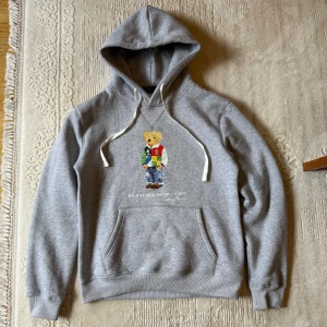 Polo Bear By Ralph Lauren Hoodie  - En klassisk hoodie i mjuk bomullsblandning med den ikoniska Polo Bear-printen på bröstet. Den grå färgen gör den lätt att matcha med allt, medan den avslappnade passformen och den bekväma huvan ger en stilren men avslappnad look. Perfekt både till vardags och när du vill ha en sportig men stilfull outfit.