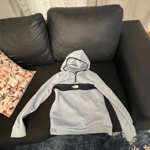 Grå hoodie från The North Face - Snygg grå hoodie från The North Face med svart detalj över bröstet och logga framtill. Tröjan har huva, lång ärm och en praktisk half zip-dragkedja. Perfekt för dig som gillar en clean och sportig stil.
