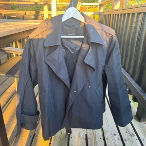 Blå  trenchcoat från Massimo Dutti - Säljer en blå trenchcoat från Massimo Dutti med klassisk krage, dubbelknäppning och snygga detaljer vid ärmsluten. Jackan har en loose passform