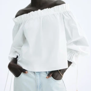 Vit offshoulder blus med volang - Superfin vit blus med offshoulder-modell och bred volangkant upptill. Blusen har långa, puffiga ärmar med dragsko vid handlederna för extra snygg detalj. Perfekt till jeans eller kjol för en trendig look.