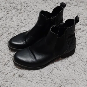Svarta Chelsea boots med glitterdetalj - Snygga svarta Chelsea boots med rund tå och platt sula. Baksidan har en glittrig detalj som ger extra edge till looken. Bootsens ovandel är i skinn och har elastiska paneler på sidorna för enkel påtagning. Perfekta för dig som gillar stilrena skor med en twist.