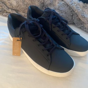 Marinblåa  - Snygga marinblåa sneakers med vit platt sula och klassisk rund tå. Skorna har snörning och är tillverkade i återvunnen polyester, vilket gör dem både stilrena och miljövänliga. Perfekta för dig som gillar minimalistisk design och vill ha ett par fräscha sneakers till vardags.