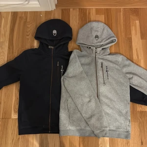 Sail Racing hoodie med dragkedja - Två snygga hoodies från Sail Racing, en i mörkblått och en i ljusgrått. Båda har huva med logga, hel dragkedja och en bröstficka med zip. Perfekt för chill dagar och har en clean, sportig look. Materialet känns mjukt och är troligen polyester eller bomull.