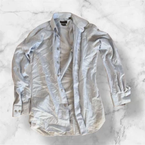 Ljusblå skjorta från Calvin Klein - Snygg ljusblå skjorta från Calvin Klein Collection i slim fit-modell. Skjortan har klassisk krage, långa ärmar och knappar framtill. Tillverkad i ett tunt, strukturerat bomullstyg som känns fräscht och stilrent. Perfekt för dig som gillar en clean och modern look.