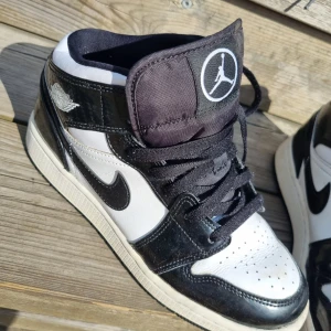 Nike Air Jordan 1 Mid svart/vit 39 - Snygga Nike Air Jordan 1 Mid i svart och vitt med klassisk Jumpman-logga på plösen. Skorna har svarta snören, svart swoosh och detaljer i blankt skinn. Perfekt för dig som gillar streetstyle och vill sticka ut med ikoniska sneakers.