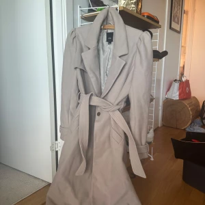 Beige kappa från River Island med bälte - Snygg beige kappa från River Island med markerade axlar, bred krage och knytbälte i midjan. Lång modell med knappar framtill och fickor på sidorna. Perfekt för dig som vill ha en stilren och trendig look under höst och vinter.Finns lite defekter på den, den är trasig i innertyget, även på knapparna och finns även en liten fläck. Inget som syns när den används