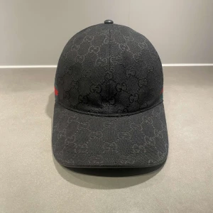 Svart gucci keps - Svart keps från Gucci med diskret GG-monogram över hela ytan och klassiska gröna och röda ränder på sidan. Justerbar rem baktill och böjd skärm. Tillverkad i bomull med exklusiv känsla och snygg passform.