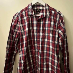 Rutig röd skjorta - Röd och vit rutig skjorta med blå detaljer. Klassisk krage, långärmad och knäppning framtill. Skjortan är i bomull och har en bröstficka. Perfekt för en avslappnad och cool stil.