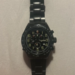 Svart kronografklocka Swiss Military - Stilren svart kronografklocka från Swiss Military Hanowa med metallarmband och tydliga siffror i gult. Urtavlan har flera små ur och tachymeter runt ringen. Perfekt för dig som gillar sportig och robust design.