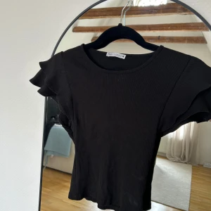 Svart ribbad topp med volangärm Zara - Svart ribbad topp från Zara med korta volangärmar och rund halsringning. Snygg croppad passform som ger en trendig vibe. Perfekt att matcha med jeans eller kjol för en cool look.