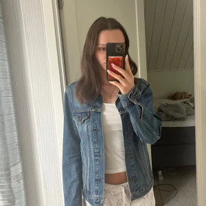 Levi's klassisk blå jeansjacka - Säljer en klassisk blå jeansjacka från Levi's med snygga detaljer och knappar framtill. Jackan har två bröstfickor med lock, raka sömmar och en tidlös passform. Mycket bra skick och sparsamt använd💙 storlek S
