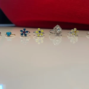Färgglada ringar med motiv, 8-pack - Säljer ett set med åtta ringar i silverfärg och en som är guldig med olika motiv: blått hjärta, blå blomma, gula blommor, glittrig diamantform, krona i rosa och grönt, samt en söt kanin. Ringarna har lekfulla färger och former, perfekta för att mixa och matcha till din stil.