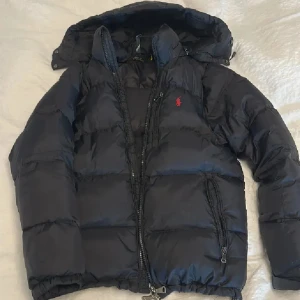 Svart dunjacka Polo Ralph Lauren M - Svart dunjacka från Polo Ralph Lauren med huva och dragkedjor framtill. Klassisk pufferstil med röd broderad logga på bröstet. Jackan har flera fickor med dragkedja och en skön, vadderad känsla som håller dig varm under kalla dagar.