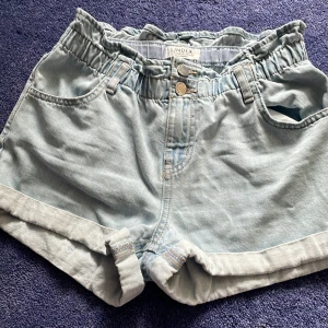 Ljusblå jeansshorts från Lindex - Snygga ljusblå jeansshorts från Lindex med elastisk hög midja och dubbla knappar framtill. Uppvikta benslut och bälteshällor. Perfekta för varma dagar och enkel att matcha med allt.