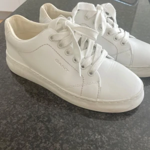Vita sneakers från Gant i skinn - Stilrena vita sneakers från Gant i skinn med klassisk design och rund tå. Skorna har snörning, diskret logga på sidan och läderdetalj bak på hälen. Perfekta för en clean och fräsch look året runt. Knappt använda!