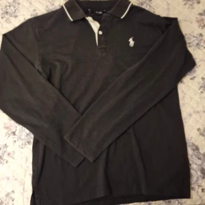 Svart långärmad pikétröja från Ralph Lauren - Klassisk svart långärmad pikétröja från Ralph Lauren med vit logga på bröstet och vit kragkant. Tröjan har knappar vid halsen och är perfekt för en stilren look. Ser ut som om den är k storleken S.