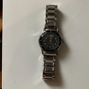 Silverfärgad Hilfiger klocka - Liten, Stilren klocka från Hilfiger med svart urtavla och klassisk logga. Klockan har en rund boett och länkarmband i silverfärgat metall. Snygga detaljer runt urtavlan och tydliga siffror gör den lätt att läsa av. Perfekt accessoar för att lyfta din outfit.