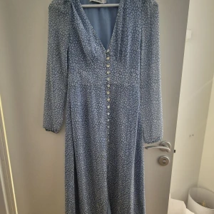 Blå mönstrad långklänning med knappar - Säljer en blå långklänning med vita tassmönster och v-ringning. Klänningen har långa ärmar och knappar hela vägen framtill. Perfekt för dig som gillar en feminin och trendig stil nu till sommarens fina dagar.