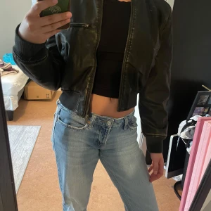 Grön/brun skinnjacka - Säljer en grön/brun skinnjacka med croppad passform och dragkedja framtill. Jackan har ribbade muddar vid ärmslut och midja samt tre fickor med dragkedja. Helt ny. Nypris 600 kr.