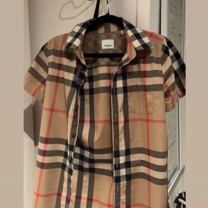 Beige kortärmad skjorta från Burberry (ÄKTA) - Säljer en  kortärmad skjorta från Burberry (ÄKTA) i beige med ikoniskt rutigt mönster. Skjortan har knappar framtill, bröstficka och har knappast används. 
