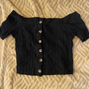 Svart ribbad offshoulder topp med knappar - Säljer en svart ribbad topp med offshoulder-design och korta ärmar. Toppen har knappar framtill och en croppad passform. Perfekt för dig som vill ha en trendig och stilren look.